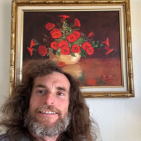 A person with long hair and a beard smiling in front of a wall with a framed painting of red flowers in a vase.